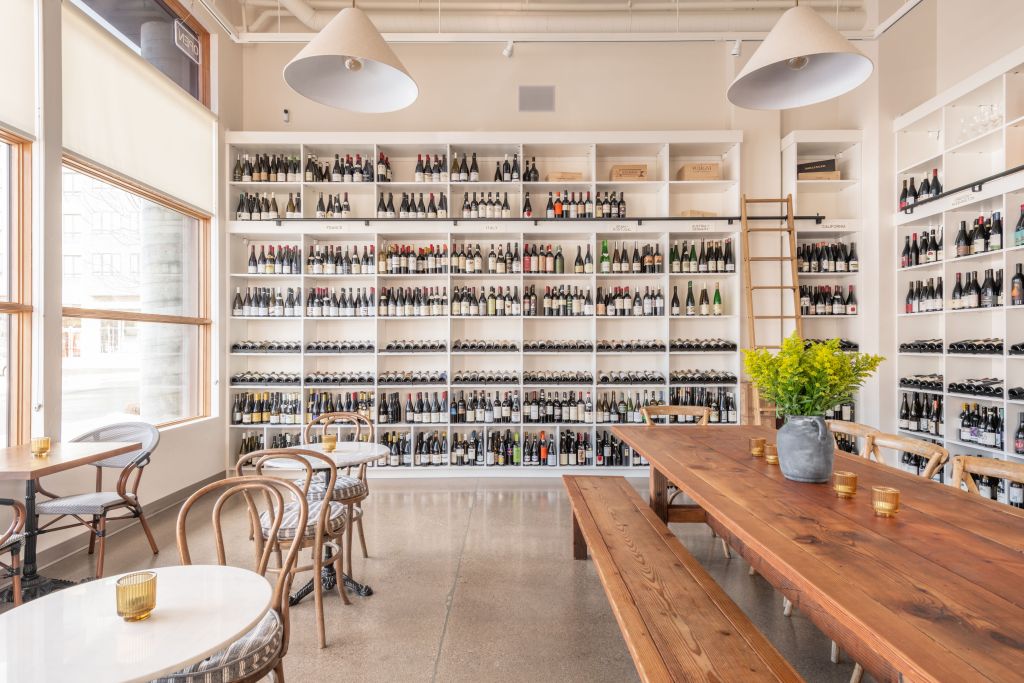Depicts a light-filled room at Viaggio Wine Merchant with two walls filled with shelves of wine and seating for patrons